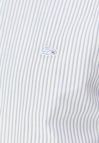 Camisa blanca con finas rayas azules, que presenta un pequeño logo bordado de cocodrilo en azul y rojo en el pecho. Tejido suave y liso.