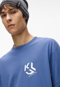 Niebieska bawełniana koszulka z okrągłym dekoltem i białym nadrukiem logo na lewej piersi. Model nosi szary dzianinowy beret z kontrastowym pasem.