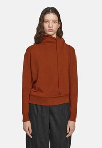 Frau mit welligem Haar, die einen rostfarbenen Pullover mit asymmetrischem hohem Kragen trägt, kombiniert mit dunkelgrauen Hosen und vor einem weißen Hintergrund steht.