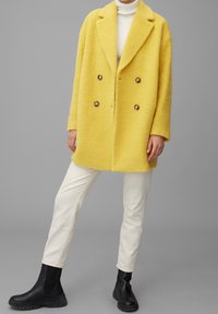 Abrigo amarillo de doble botonadura con una tela texturizada, grandes solapas y botones redondos. Combinado con pantalones blancos y botas de tobillo negras.
