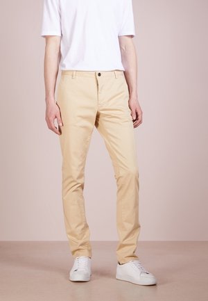 Chinos beige in cotone con vestibilità dritta, dotati di chiusura con bottone frontale, passanti per cintura e tasche laterali, indossati con una maglietta bianca e sneakers bianche.