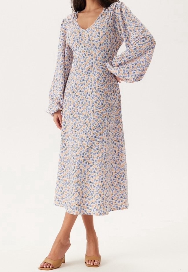 Femme portant une robe florale à manches longues, mi-longue, avec des manches bouffantes et des talons ouverts beiges, debout avec une main sur la hanche.