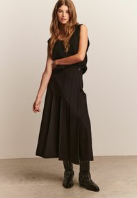 Frau mit langen braunen Haaren, die ein schwarzes ärmelloses Strickoberteil, einen weiten schwarzen Midirock und schwarze Lederstiefeletten mit Riemen trägt.