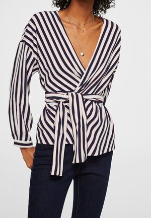 Vrouw die een marineblauwe en witte blouse met verticale strepen draagt, met een diepe V-hals en een gestrikte taille, gecombineerd met donkerblauwe spijkerbroeken.