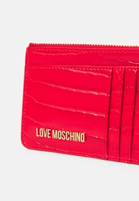 Červená lesklá peněženka z umělé kůže s krokodýlí texturou, se zlatým logem "LOVE MOSCHINO" a několika sloty na karty. Zapínání na zip.
