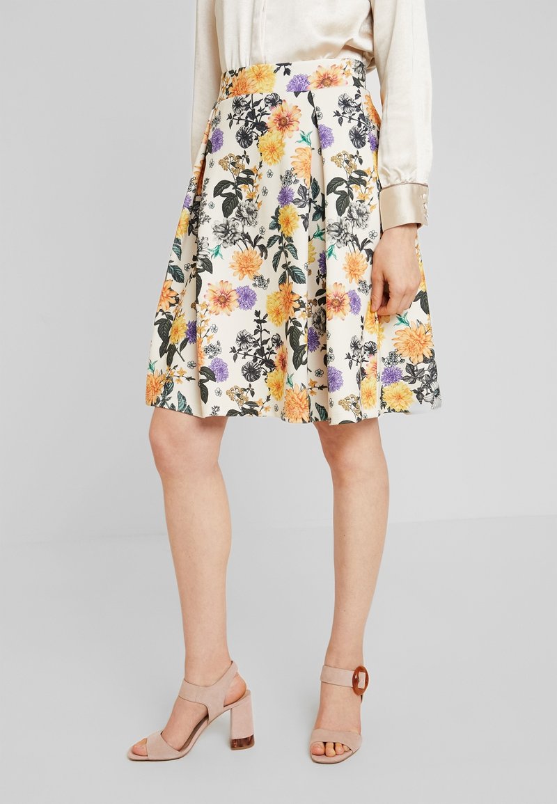 Femme portant une jupe blanche longueur genou avec imprimé floral jaune, violet et noir, un chemisier beige et des sandales beiges à talon bloc et bout ouvert.