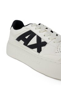 Sneaker in pelle bianca con logo "AX" nero, design con lacci e dettagli testurizzati. Presenta una suola bianca spessa con il nome del marchio impresso.