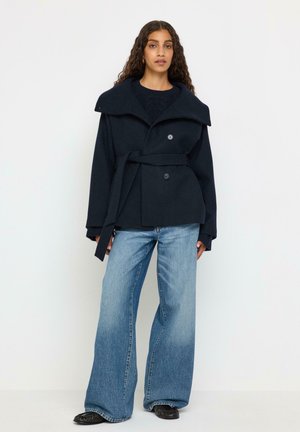 Vrouw met lang krullend haar, gekleed in een marineblauwe mantel met riem, wijde blauwe jeans en zwarte schoenen, staand tegen een effen witte achtergrond.