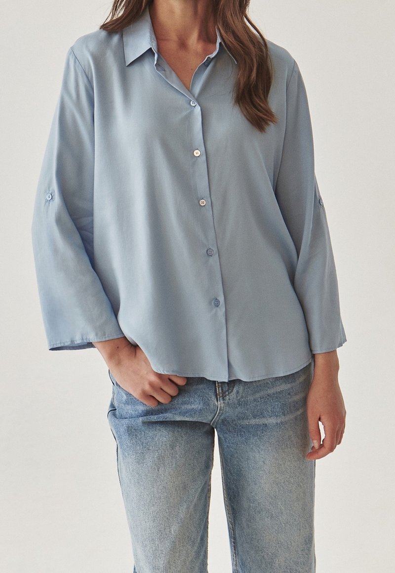 Femme portant une chemise ample bleu clair à boutons avec des manches longues et un jean en denim bleu clair, debout avec une main dans la poche.