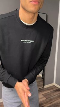Άτομο που φοράει μαύρο φούτερ με το κείμενο "WEEKEND OFFENDER ADMIT NOTHING", τρίβει τα χέρια, στέκεται σε δωμάτιο με ξύλινο πάτωμα.