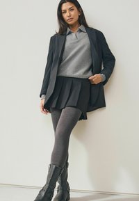 Black blazer over a gray sweater, paired with a black pleated skirt and gray tights. She wears knee-high black boots with buckles.