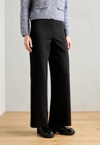 Černé kalhoty s širokými nohavicemi vyrobené z měkkého materiálu. Design zahrnuje vysoký pas a rovný střih, doplněný o mohutné černé boty.