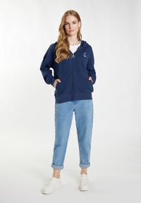 Felpa con zip di colore blu navy, con tasche frontali e ricamo a forma di ancora sul petto. Abbinata a jeans azzurri arrotolati e sneakers bianche.