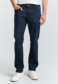 Hombre con jeans azul oscuro, una mano en el bolsillo y zapatillas blancas, de pie contra un fondo lisos claro.