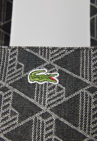 Tkanina s crno-bijelim uzorkom ima vezeni logo krokodila u zelenoj i crvenoj boji na teksturiranoj površini, naglašavajući geometrijske dizajne.