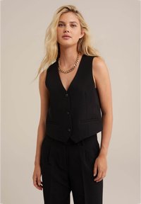 Juodas liemenė iš tekstūruotos medžiagos, su V formos dekolte, trimis priekiniais mygtukais ir dviem šoninėmis kišenėmis. Suderinta su juodomis kelnėmis.