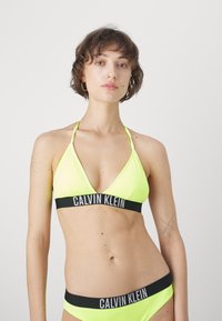Neon žlutý bikini set s trojúhelníkovým topem s tenkými ramínky a černým pásem s nápisem "CALVIN KLEIN"; hladká textura.