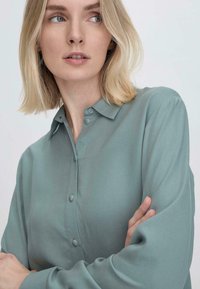 Camicia button-up verde chiaro con una texture morbida e liscia, caratterizzata da un colletto classico e maniche lunghe, che mostra una vestibilità rilassata e una leggera lucentezza.