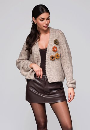 Kvinna i en beige stickad cardigan med fyra färgglada blommor, en mörkbrun linne, läderminikjol och genomskinliga svarta strumpbyxor.