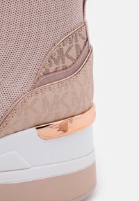 Gros plan du talon arrière d'une basket beige et rose avec des textures en tissu et cuir, un accent métallique en or rose, et un détail de motif "MK".