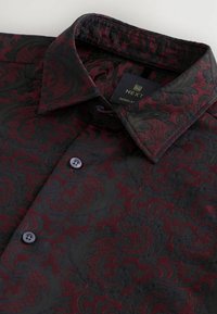 Bordeaux- og sort mønstret skjorte med paisley-design, som har en krave med knaplukning og blå knapdetaljer. Stoffet er struktureret.