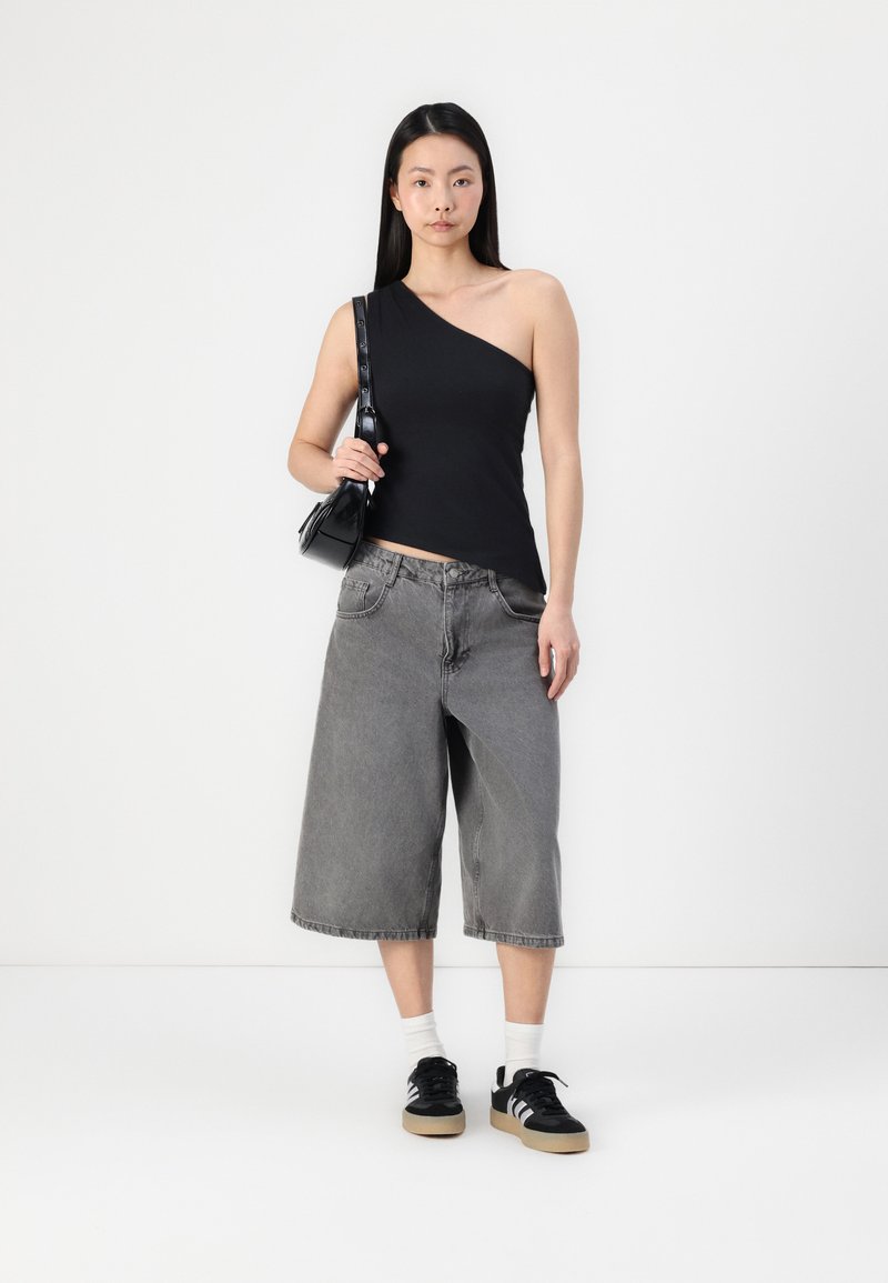 Schwarzes One-Shoulder-Top kombiniert mit grauen Denim-Culottes. Das Model trägt schwarze Sneaker und hält eine kleine schwarze Tasche. Weißer Hintergrund.