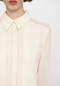 Camisa de color crema, de manga larga, con cuello puntiagudo, que presenta pliegues verticales y una tapeta de botones en la parte delantera.