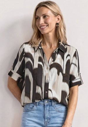 Kobieta o blond włosach, ubrana w czarno-biało-beżową, wzorzystą, krótkorękawową koszulę z guzikami wpuszczoną w jasnoniebieskie jeansy, uśmiechająca się i patrząca w prawo.