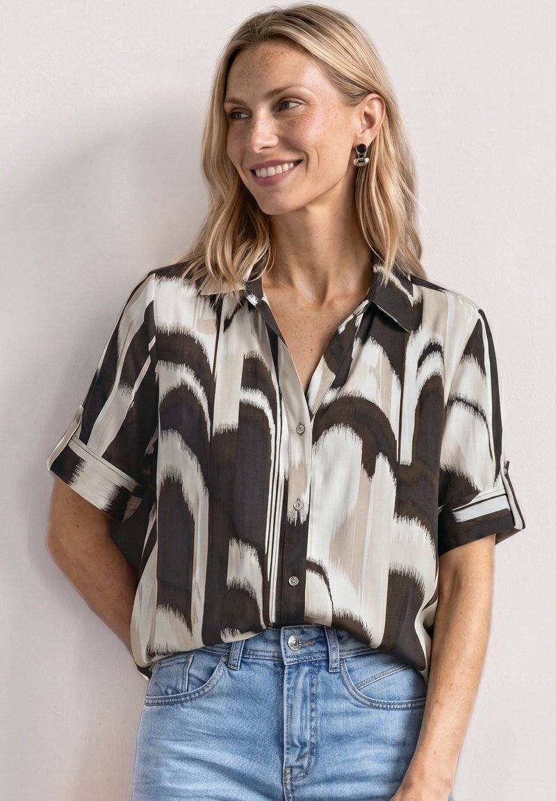 Femme aux cheveux blonds portant une chemise à manches courtes à motifs noirs, blancs et beiges, rentrée dans un jean bleu clair, souriante et regardant vers la droite.