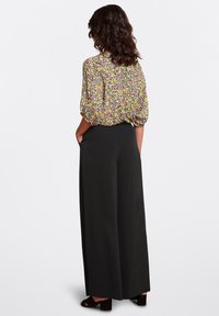 Blusa floreale con maniche a sbuffo, caratterizzata da un mix di fiori gialli e rosa, abbinata a pantaloni neri a gamba larga e sandali con tacco quadrato.