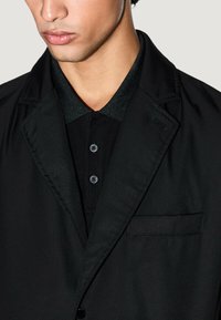 Blazer negro con un cuello verde texturizado, con un bolsillo en el pecho y dos botones, superpuesto a una camiseta polo negra.