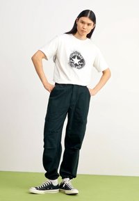 Camiseta blanca de manga corta con un logo negro de Converse, combinada con pantalones oscuros verdes de cargo y zapatillas negras con suelas blancas.