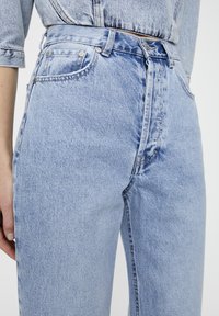 Jeans de tiro alto en azul claro con un corte recto, que presentan herrajes metálicos plateados, bolsillos clásicos y una textura de denim desgastado.