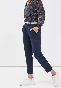 Personne en pantalon bleu marine avec ourlets roulés, ceinture blanche, haut à manches longues floral, mains dans les poches, et baskets blanches marchant sur fond blanc.