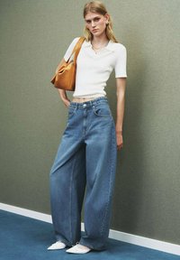 Giovane donna che indossa una maglietta bianca a maniche corte, jeans blu a gamba larga, scarpe bianche a punta, mentre porta una borsa marrone chiaro a tracolla contro un muro grigio.