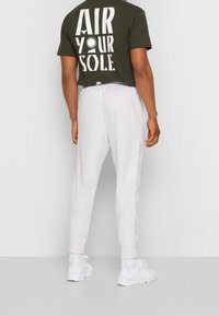 Zelené tričko s velkým bílým nápisem „AIR YOUR SOLE“ na zádech; doplněné bílými tepláky s postranními pruhy; sportovní design.