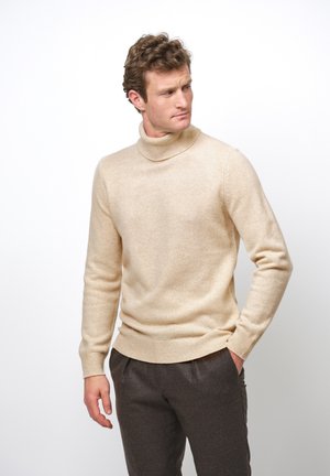 Pull en col roulé beige en tissu doux, avec des manches longues, des poignets côtelés et une coupe ajustée. Assorti à un pantalon gris foncé.