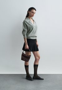 Maglione grigio chiaro lavorato a maglia con scollo a V, gonna nera a campana, calze nere e ballerine. Tiene una piccola borsa bordeaux.