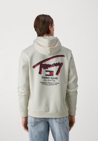 Lichtbeige hoodie met grafische print in rood en zwart. Heeft een groot "Tommy" logo en stadsnamen op de achterkant. Gemaakt van een katoenmix.
