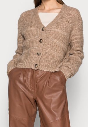 Frau trägt eine strukturierte beige Strickjacke mit Knöpfen über einem weißen Oberteil, kombiniert mit hoch taillierten braunen Lederhosen, Hände in den Taschen.