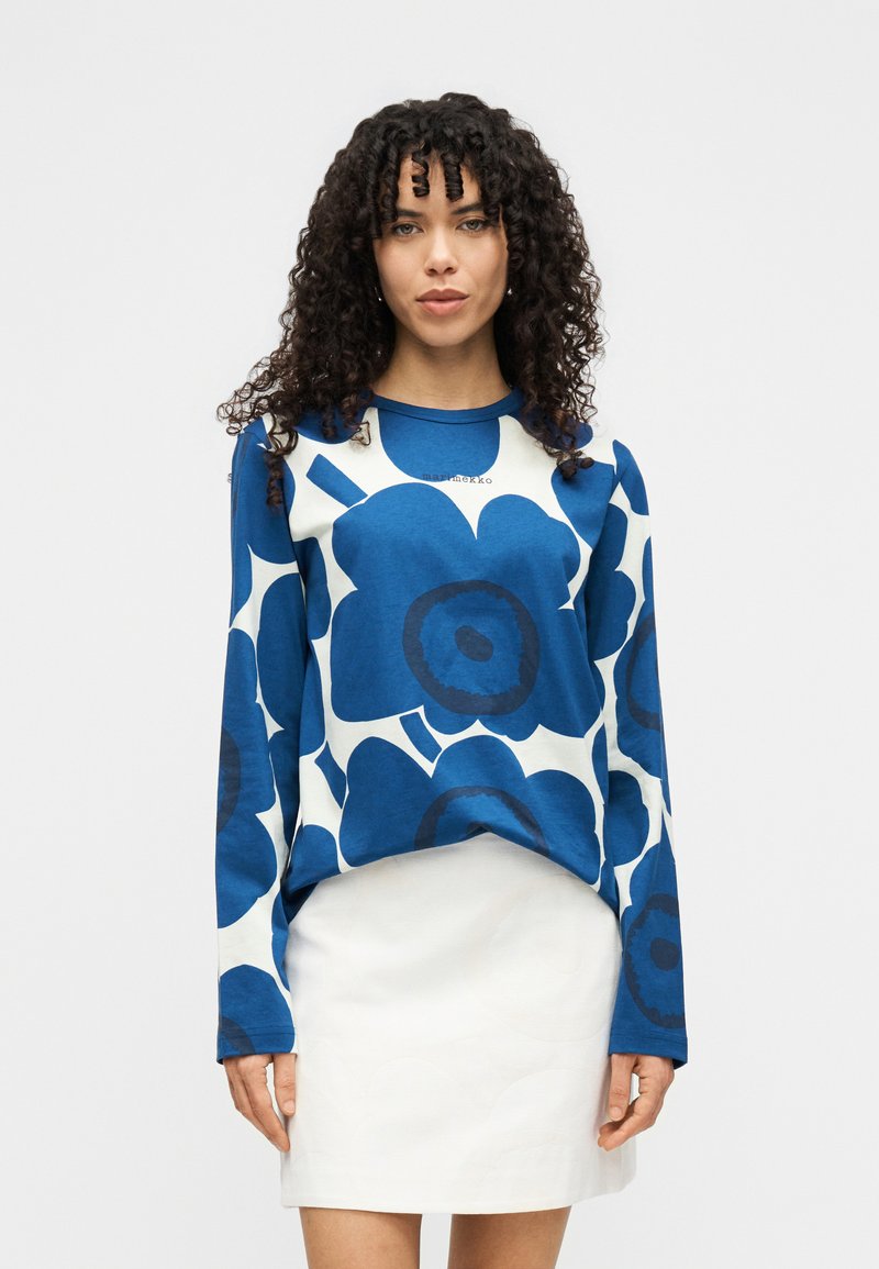 Μπλε μπλούζα με μακριά μανίκια και μεγάλο λουλουδάτο μοτίβο σε σκούρο μπλε και λευκό, με στρογγυλή λαιμόκοψη, συνδυασμένη με λευκή φούστα. Μαλακή υφή υφάσματος.