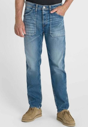Uomo che indossa jeans blu medio a gamba dritta con grandi tasche applicate frontali e scarpe beige in camoscio, una mano nella tasca.