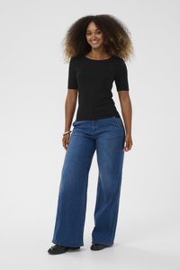 Femeie zâmbitoare cu păr creț, îmbrăcată cu un top negru cu mânecă scurtă, blugi largi albaștri și pantofi negri, stând în fața unei fundal alb.