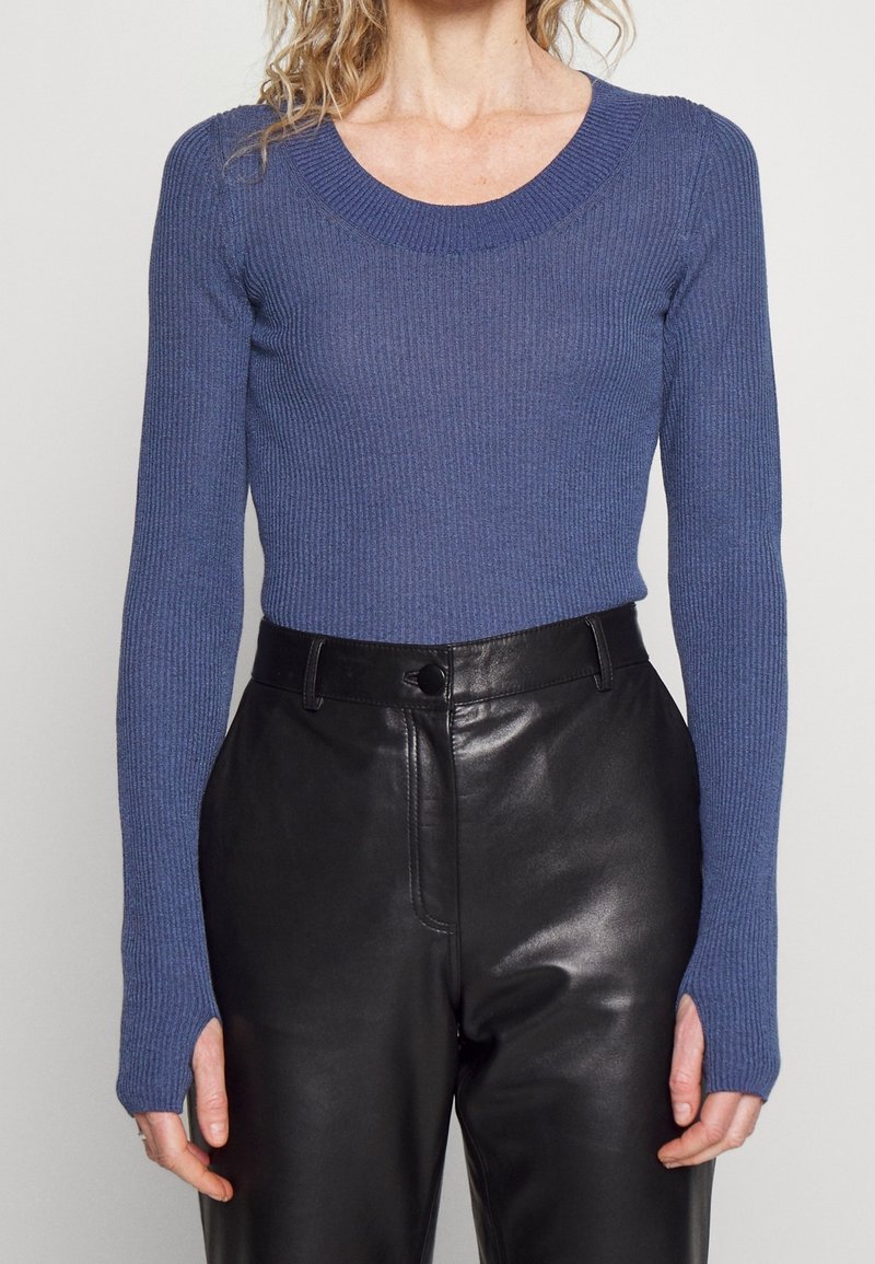 Femme portant un haut bleu moulant à manches longues côtelé avec des trous pour les pouces, associé à un pantalon en cuir noir taille haute.
