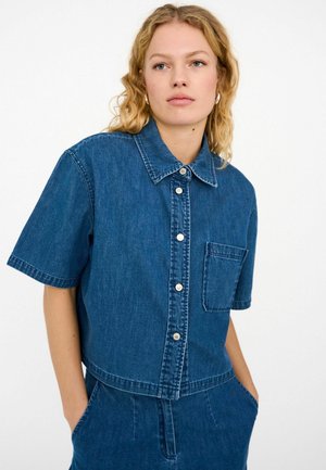 Žena s kučeravými blond vlasmi, oblečená v modrej rifľovej košeli s krátkym rukávom a zapínaním na gombíky, so zapínacím vreckom vpredu, a v zladených modrých rifľových nohaviciach, s rukami v vreckách.