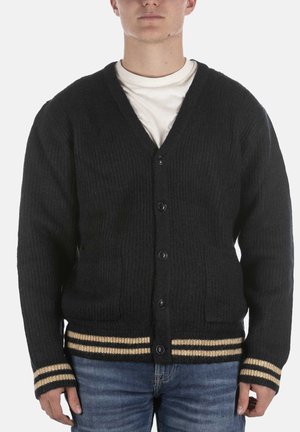 Man draagt een zwarte gebuttonde cardigan met beige strepen op de manchetten en zoom over een wit overhemd en blauwe spijkerbroek.