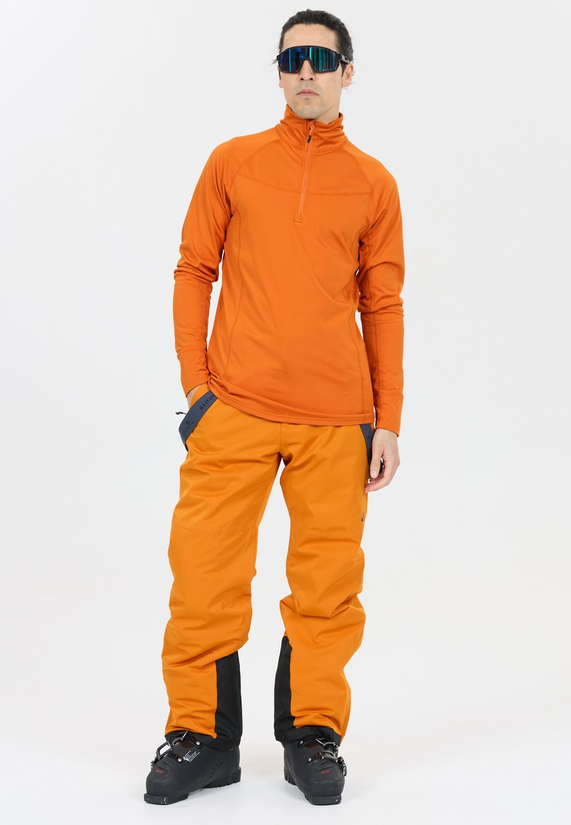 Oranssi urheilullinen pitkähiha toppi puolivetoketjulla ja oranssit lumihousut mustilla yksityiskohdilla, yhdistettynä mustiin hiihtokengiin.