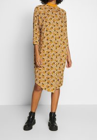 Hnědé šaty s vzorem, tříčtvrtečním rukávem, zdobené květinovými a abstraktními vzory. Okraglový výstřih a asymetrický lem, doplněné černými kotníkovými botami.