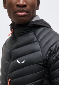 Κοντινό πλάνο ενός ατόμου που φοράει μαύρο μπουφάν με κουκούλα και πορτοκαλί κορδόνια φερμουάρ, με λευκό γωνιώδες λογότυπο στο στήθος.