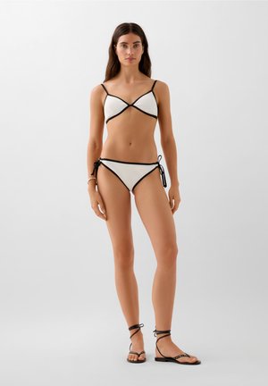White ribbed bikini with black trim, featuring a triangle top and side-tie bottom. Paired with black flat sandals with ankle straps.